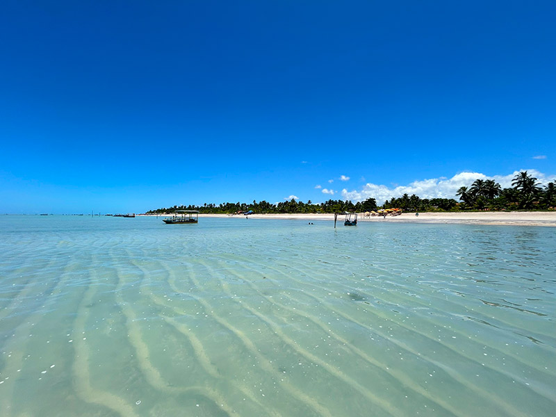 Mar vazio e cristalino com barcos ao fundo e céu azul na Praia de Antunes