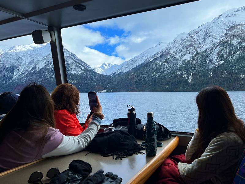 Pessoas tiram fotos com celular de montanha com neve em Bariloche a partir de barco panorâmico em lago