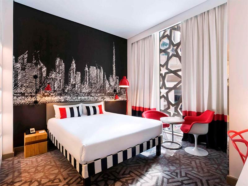 Quarto do Ibis Styles Airport Dubai com arte da cidade ao fundo, cama de casal e decoração em preto, branco e vermelho